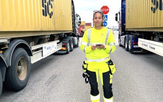 Ung kvinna i gula reflexkläder står mellan två conainerbilar.