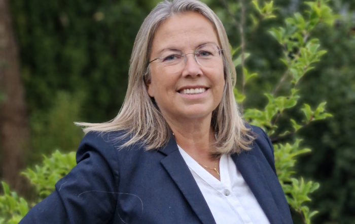 Eva Lindell, ny vd Norrköpings Hamn, kvinna, leende, i blå kavaj, grön växtlighet i bakgrunden.