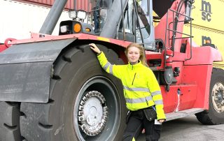 Ung kvinna i gula reflexkläder lutar sig mot det stora hjulet på en containertruck som står parkerad.