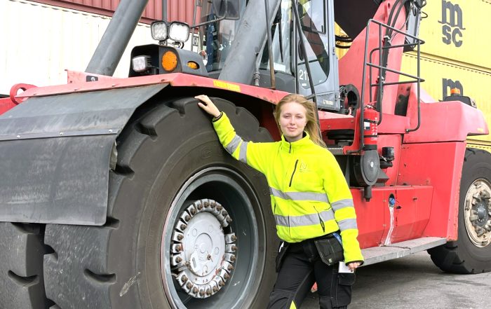 Ung kvinna i gula reflexkläder lutar sig mot det stora hjulet på en containertruck som står parkerad.