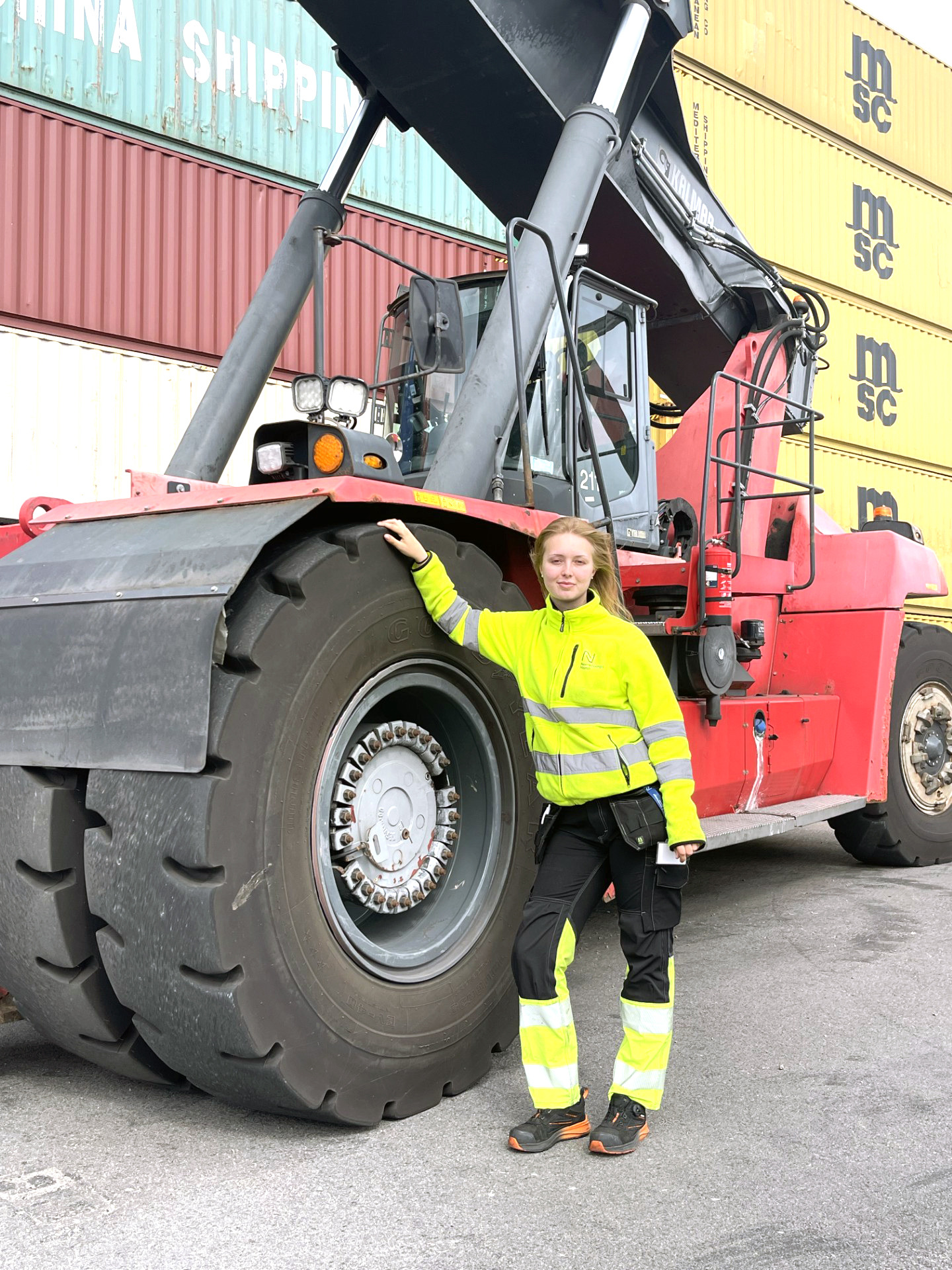 Hedda Ung kvinna i gula reflexkläder lutar sig mot det stora hjulet på en containertruck som står parkerad.
