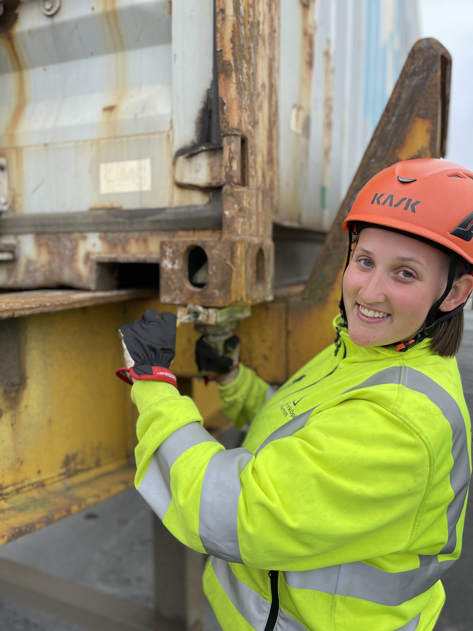 Heidi Leende ung kvinna i gula reflexkläder och orange skyddshjälm vid container.