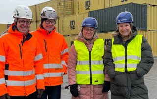 Två män i orange refelxjackor och vita hjälmar, två kvinnor i gula reflexvästar och blå hjälmar. I bakgrunden containrar,