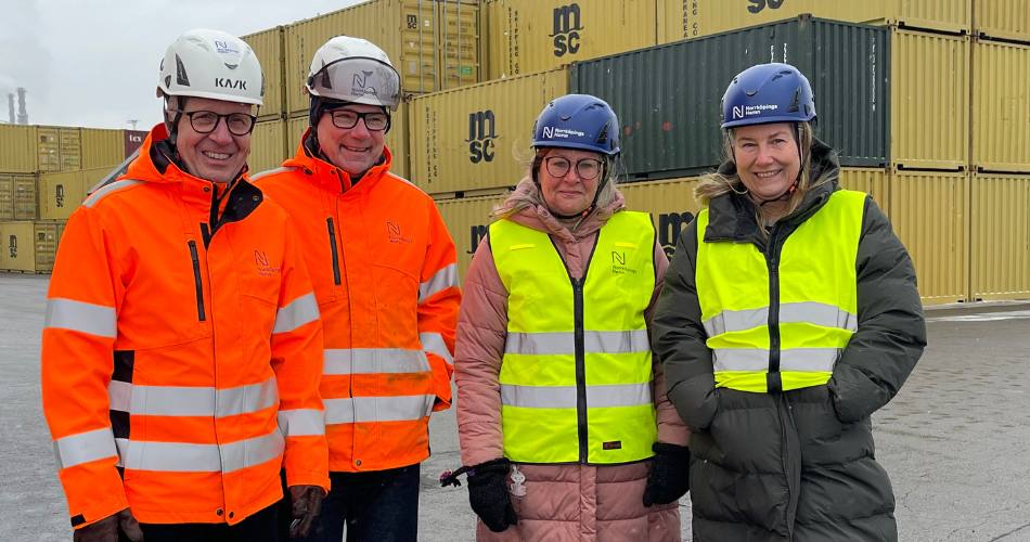 Två män i orange refelxjackor och vita hjälmar, två kvinnor i gula reflexvästar och blå hjälmar. I bakgrunden containrar,