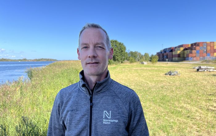 M Malzoff, man i grå tröja, mot bakgrund av vatten, grönska och containrar.