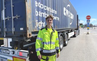 Leende ung man i gula reflexkläder står framför en lastbil med en blår container.