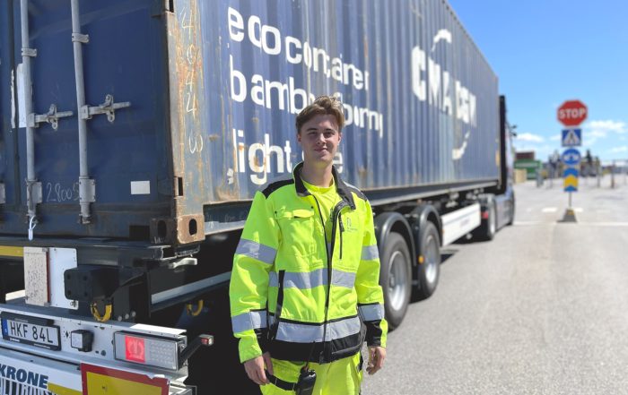 Leende ung man i gula reflexkläder står framför en lastbil med en blår container.